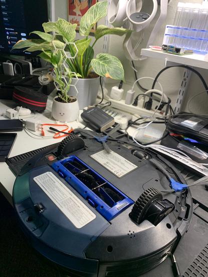 A vacuum bot on a repair station, with plants and wires in the background
