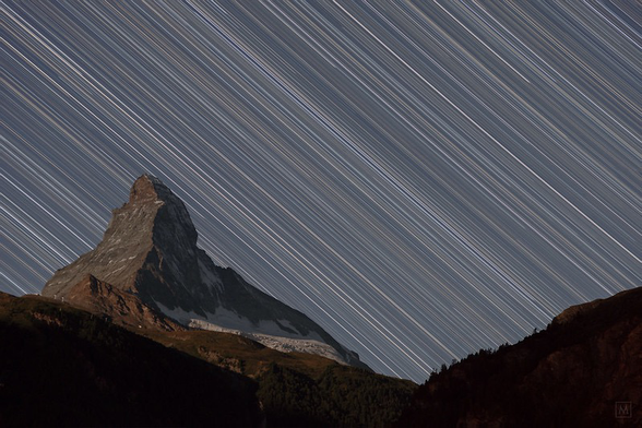 The Matterhorn stands in contrast to star trails that streak from upper left to lower right. The sky is a charcoal blue color as the moon is rising behind the photographer gently illuminating the face of the Matterhorn.