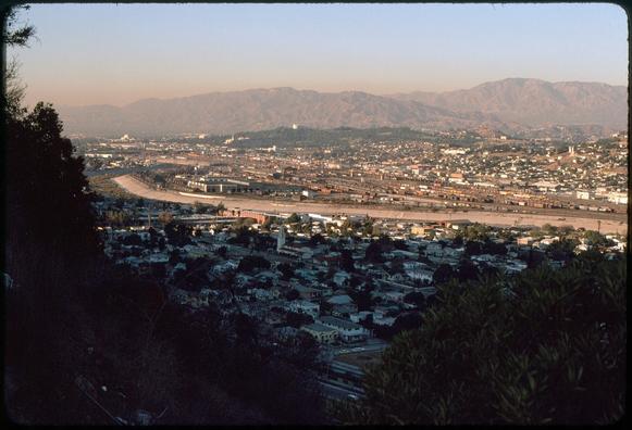 A view across an urban valley toward a ridge of mountains in the distance. In the foreground is a residential area. Beyond, the concrete embankments of the Los Angeles river Slice across the frame. A large railyard and shops are beyond the river. In the middle distance a wooded hillside marks the location of Forest Lawn Cemetery.