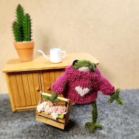 A small knitted frog is wearing a pink sweater with a white heart. He is carrying a box of pink and white flowers. On a table behind him is a pot with three small cacti in it and a white watering can.