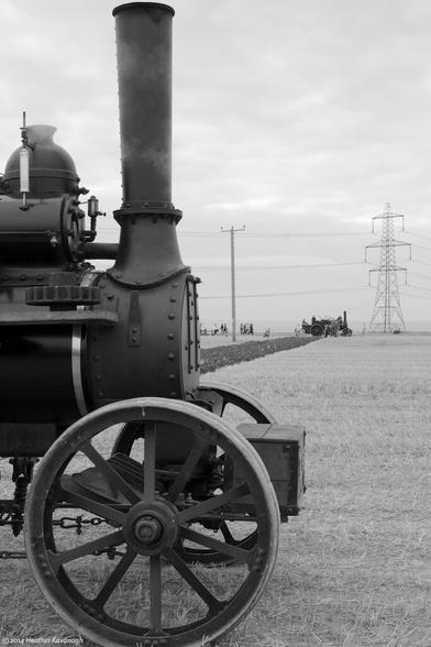 A huge twelve ton steam traction engine dominates the scene. In the background is the other engine. Between them, hauled by a steel cable wound round powered drums beneath each engine, is a reversible plough.