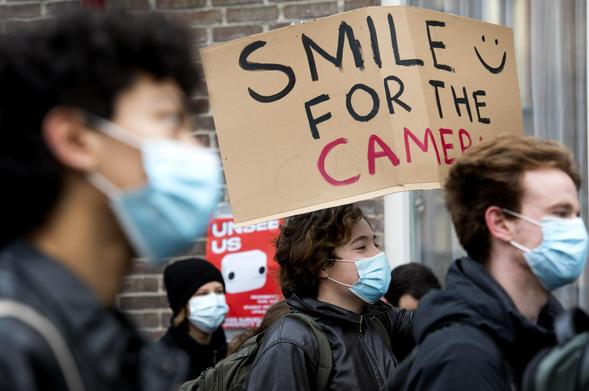A photo of a protest against the use of smart camera's in Leiden University. One protester holds a sign that reads 'Smile for the Camera'