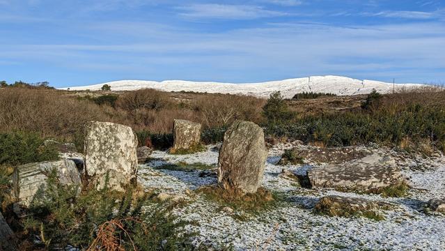 A stone circle in strong light against reddish-purple bare trees and a distant ridge covered in fresh snow, a blue sky above with thin whispy clouds.
