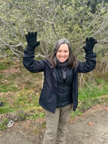 Catherine dressed with hiking pants, a black jacket, and scarf, her arms raised, and pretending to be a ferocious black bear. Forest in the background.