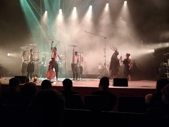 Danish-Norwegian-German folk band Heilung performing at the Bridgewater Hall in Manchester. A group of performers wearing pantaloons and holding round shields aloft stand to the left of the stage. A man wearing a headdress with antlers and a large robe stands palm to palm with a woman in Romano-British dress to the right of the stage.