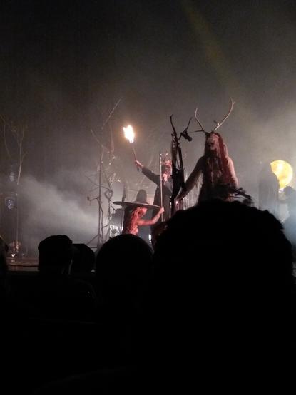 Danish-Norwegian-German folk band Heilung performing at the Bridgewater Hall in Manchester. A woman in a headdress with antlers stands at a microphone right of centre. Slightly behind her to the left of centre a man in a headdress with antlers hits the top of a round shield with a flaming torch. The shield is supported by a woman wearing pantaloons, her upper body naked and tattooed.