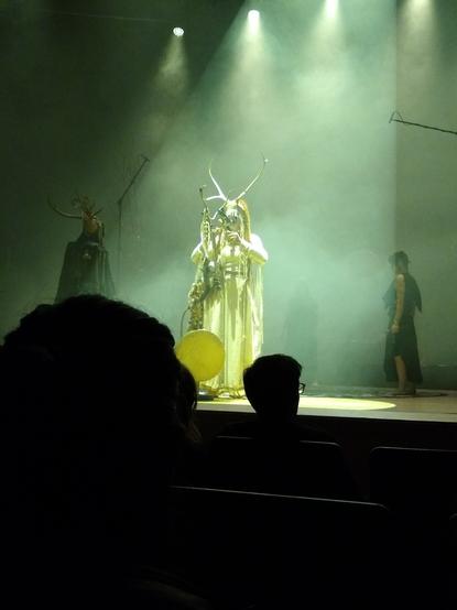 Danish-Norwegian-German folk band Heilung performing at the Bridgewater Hall in Manchester. A woman in a headdress with antlers stands at a microphone hitting two small bones against each other. To the left stands a man in a headdress with antlers, his back to the audience. To the right a woman in Romano-British dress is walking towards the centre of the stage.
