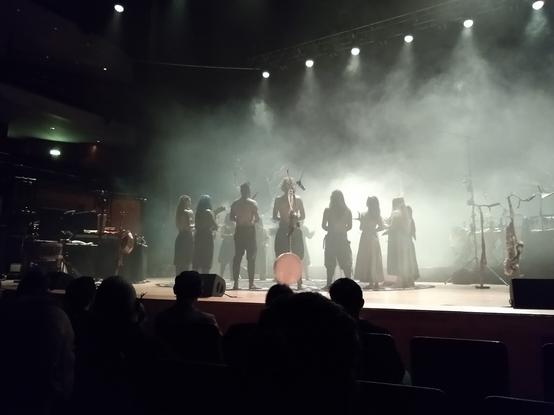 Danish-Norwegian-German folk band Heilung performing at the Bridgewater Hall in Manchester. A group of people stand in a circle, facing inwards. They are being cleansed with incense by a man in the centre of the circle. Each person carries an animal bone or antler.