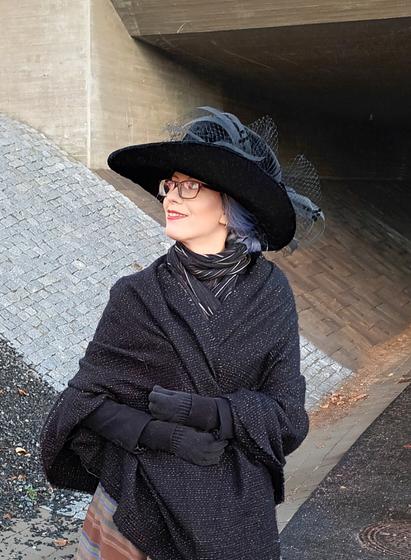 A digital crop (which is to say it doesn't have very many pixels) of Sini Tuulia outside, wrapped in a black and grey shawl. She is wearing an enormous black velvet hat with a wide, partially upturned brim. It is as wide as her shoulders and adorned with feather-like flat crinoline tube bits, black dotted veiling and artful folds of the velvet. The hat looks like it would look slightly ridiculous on its own, but with everything else Sini is wearing, it sort of makes sense.
