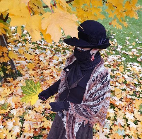 A photo of Sini looking at a half green and half yellow maple leaf, one of hundreds littered on the ground at her feet and on the tree just in frame. The angle is such that you can see the decorations of her much smaller hat. It has a rounded crown, a narrow brim that is turned up at one side, a velvet hat band, petersham rosettes and black netting. She is also wearing a black fabric face mask on top of a white one, has a little lapel pin, red orange butterfly wing earrings and a brown and tan crochet shawl.