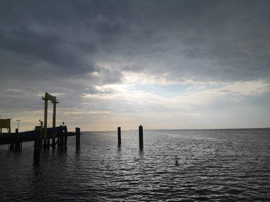 Am Himmel eine abziehende Gewitterwolke, am Horizont zeigt sich die Sonne wieder, davor als schwarzer Scherenschnitt der Fähranleger mit dem Gezeitenhebewerk für die Anlegerampe