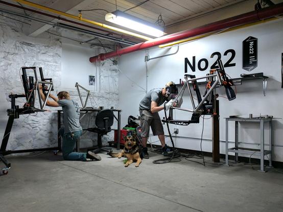 Rahmenbau bei 22 Bicycles. 2 Personen schweißen Titanrahmen, dazwischen liegt ein Schäferhund.