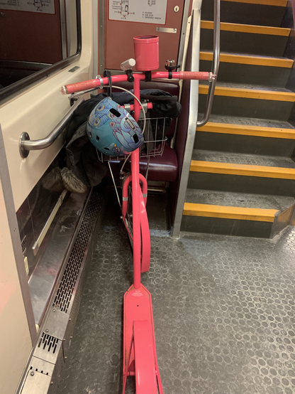 A neon pink kickbike nestled on an MBTA commuter train, with a peacock-adorned helmet dangled from the handlebars