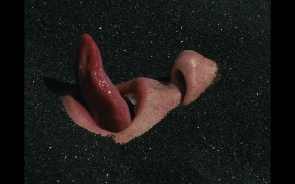 A face half buried in black sands with a tongue sticking out.
