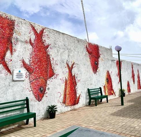 Streetartwall. On a long gray wall are sprayed several (8-10) large red fish, head down. Between them are green park benches. It is part of an exhibition of the artist in adjacent building.