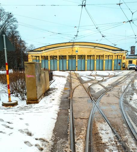 Old yellow tram depot building with multiple curving rail tracks leading to it and footprints in the snow leading to a park off to the left