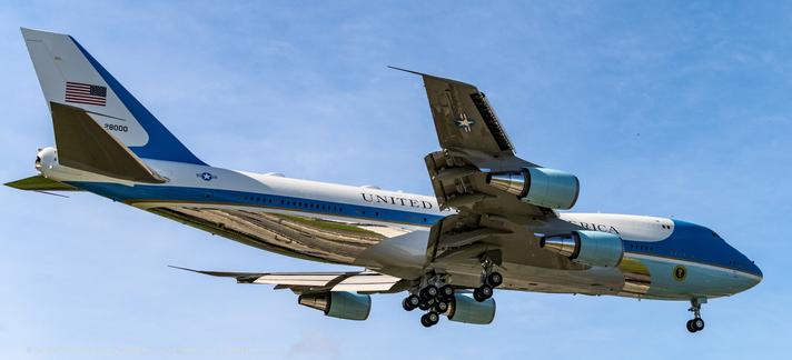 a large 4 engine aircraft with its landing gear down as well as its flaps, floats in to land. The plane is white with blue accents and says 'United States Of America" in large letting on the fuselage. The lower part of the plane is shiny polished aluminum.