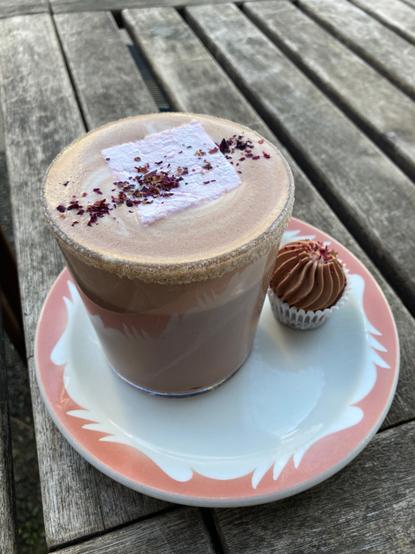 A slightly further away shot of the hot chocolate; a tiny pink chocolate cupcake with pink icing sits beside it on the saucer.