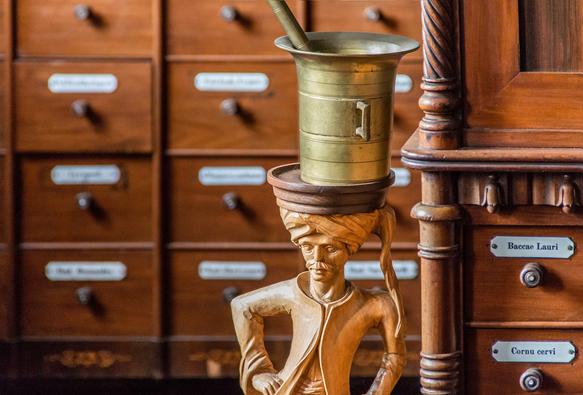 Wooden stand carved in the shape of a standing man with a turban and a mustache. On top of the stand is a copper looking crucible with some instruments handles protruding. In the background is cabinet of drawers.