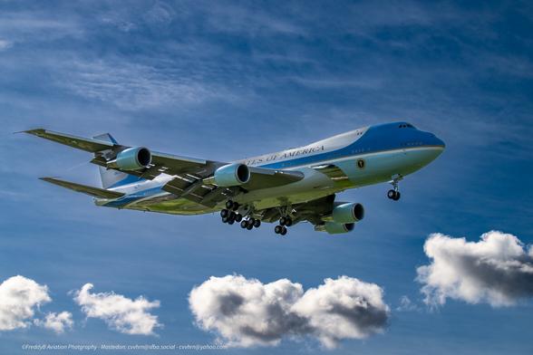a large 4 engine plane floats in for a landing. Its gear is lowered and the plane is light blue and polished aluminum at the base with a blue coclpit area swooping back to the rear. The rest of the plane is white. ,United States of America is emblazoned on the fuselage. Just behind the front gear and on the side the round seat of the president