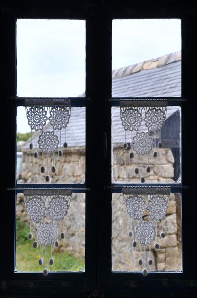 Blick von innen durch ein Sprossenfenster mit hellblaue Rahmen auf ein Scheune aus Naturbruchsteinen mit Schieferdach. In den 4 unteren Fensterfeldern hängen jeweils drei zu einem Dreieck angeordnete gehäkelte Schmuckrosetten mit 5 angehängten ebenfalls gehäkelten Pompons