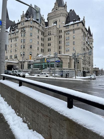 The Chateau Laurier hotel in downtown Ottawa, it has a gothic sort of look. The road in front of it is wet and there is snow.