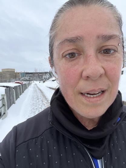 Me, a white cod woman runner, wearing a black jacket & buff. Behind me is a snow covered path.