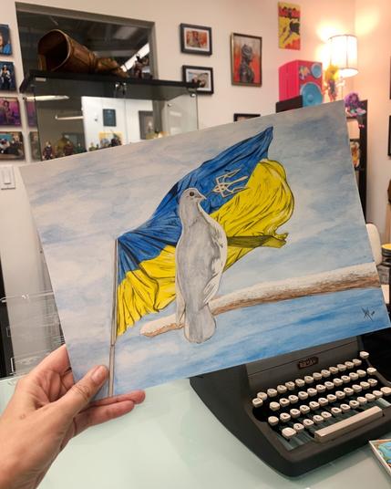 A hand holds a water colour painting of a dove perched on a branch in front of a Ukrainian flag. The dove is holding a Tryzub in its beak.