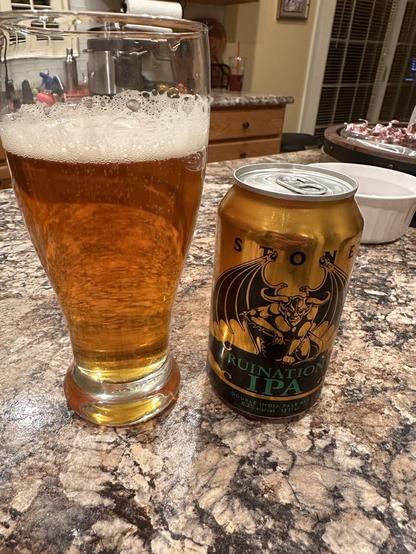 A 12 oz can of Stone Ruination IPA beside a glass of orange-amber beer