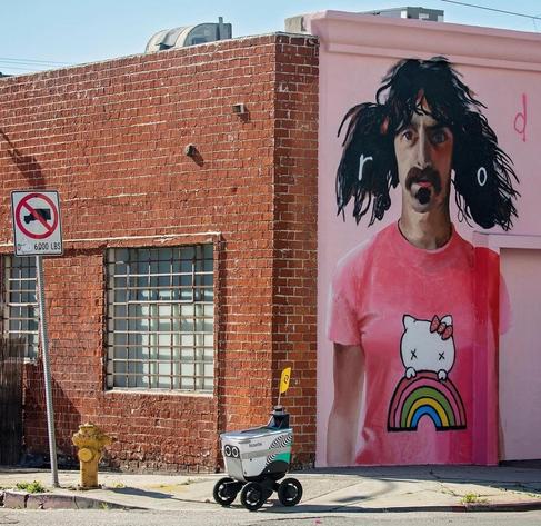 Streetartwall. A mural on an exterior wall of a red brick building, a corner house whose one side was primed pink. Depicted is a mustachioed man with braids and pink Hello Kitty shirt. It is the musician Frank Zappa. The photo is further enhanced by a @serverobotics robot named "Noelle", (who was slowly driving by to deliver his next package).