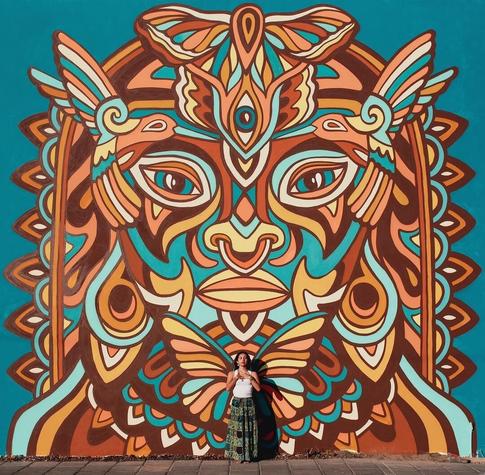 Streetartwall. A mural in the style of indigenous wall painting. In the colors turquoise, brown, yellow and white shapes, arcs and lines are connected to form a mask, which shows a kind of animal-human head with earrings, nose ring and headdress. Under the mouth is a butterfly in front of which a real woman for the photo.