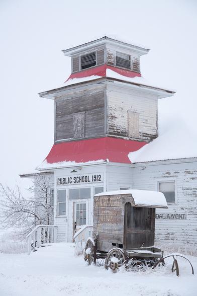 Winter scene with deep snow. A very ancient wooden-wheeled wagon (wild west style) stands in the snow outside the front of a long abandoned school house. The schoolhouse, with its peeling paint and wooden boards, is dated 1912 and ceased use decades ago.