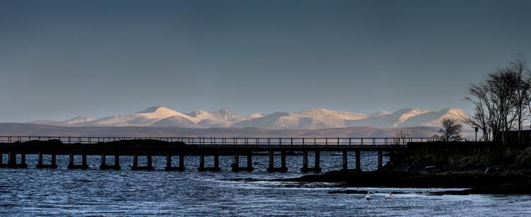 Panoramic photograph of the 'still' snow covered peaks on the Isle of Arran, a fair distance from the harbour where I'm standing. Metered for Arran so the foreground is in silhouette showing a stretch of the bay and the commercial pier.