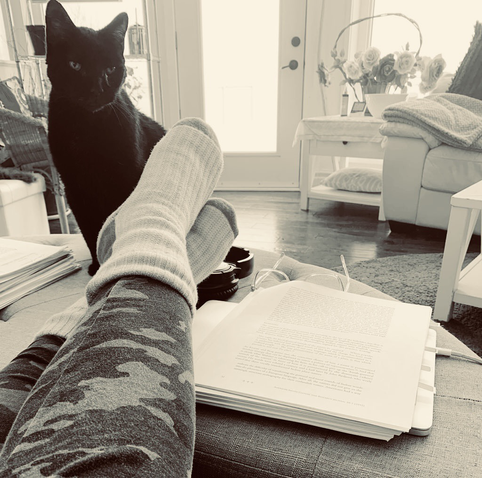 This is a photo of the author’s legs stretched out on a living room ottoman. On my feet are a pair of heavy knit socks. Beside my feet sits a black cat. To the right of my feet are a stack of printed papers. The photo is in black and white with a slight sepia tone.