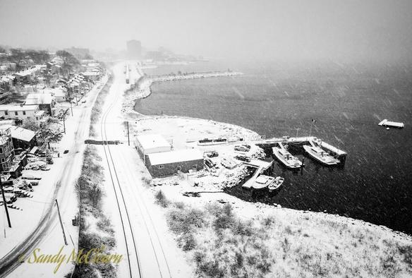 Black and white landscape photo taken at height along the waterfront with the harbour at right and road and train tracks and houses at left.