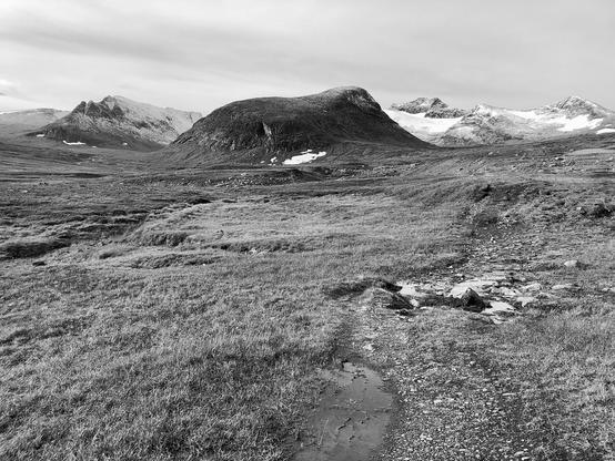 Schwedisches Fjällstation. Vorne Wiese, Steine und ein kleiner Bach. Am Horizont in der Bildmitte eine sehr großer, rund geformter Berg, dahinter schneebedeckte Grate und Gipfel. Bewölkter Himmel