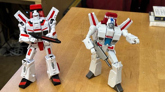 Two toy robots next to each other. On the left, NewAge Firebird in Jetfire's original toy colours, a white, winged robot with black and red detailing that makes the Macross inspiration obvious. On the right, Magic Square Meteorite, which has mostly the same silhouette but in a colour scheme much closer to Jetfire/Skyfire's appearance in the Transformers cartoon.