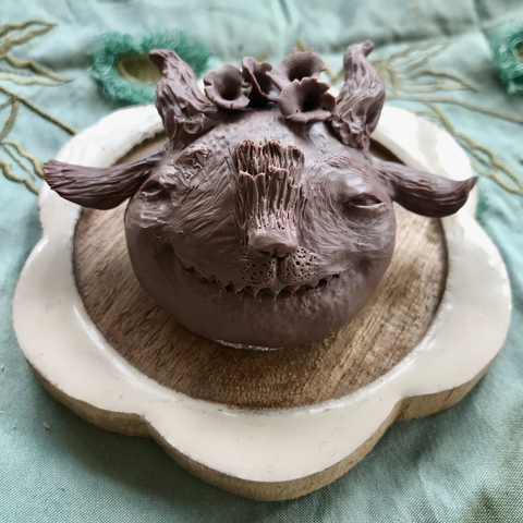 Small sculpt. Brown Cosclay. Goatee ears, pointy teeth, flat hairy nose, horns. Smirking because it has flowers tangled in its fur.