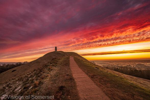 A bright red sky over Glastonbury Tor. A pathway leads up the tower on top of the hill.