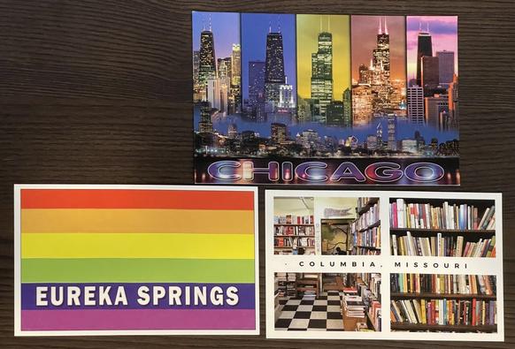 Three postcards: the top postcard is five different multi views of Chicago skyline Towers, including John Hancock and the tower formerly known as the seers tower and pretty colors at the bottom of the postcard. It reads Chicago. The bottom left postcard is a gay pride flag with the words Eureka Springs on it, the bottom right postcard is a multi view of a bookstore from Columbia Missouri books line the shelves and the floor is a black and white checkered pattern