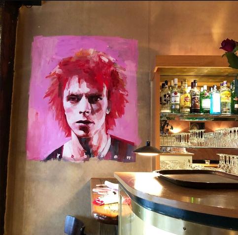 Street Art. In a pub in Berlin is this small mural painted by David Bowie. It is on a pink background. Bowie has red tousled hair and looks at us. Next to it is a bar.
Info: The pub "Neues Ufer" (Double meaning: New Shores/New start) 
was one of the first gay pubs in Berlin. Bowie lived a few houses away, together with Iggy Pop.