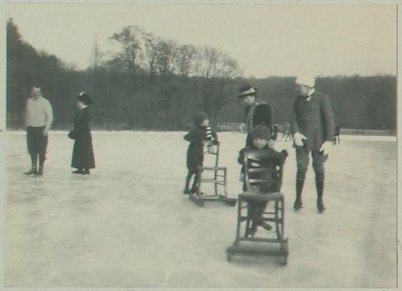 Schwarz-weiß Fotografie, mehrere Menschen mit Schlittschuhen auf einer Eisfläche, zwei Jungen nutzen Stuhlschlitten als Stütze