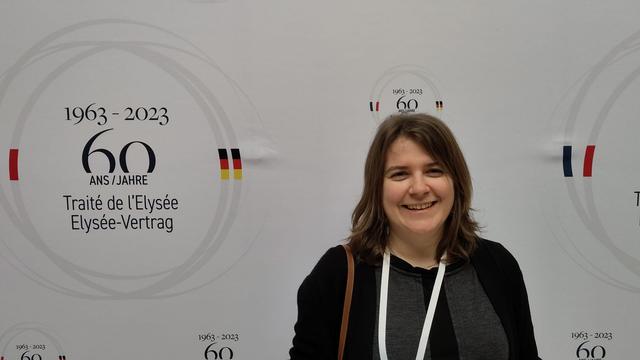 Woman standing in front of a white wall with the inscription "1963 - 2023, 60 years of the Elysee Treaty" in German and French