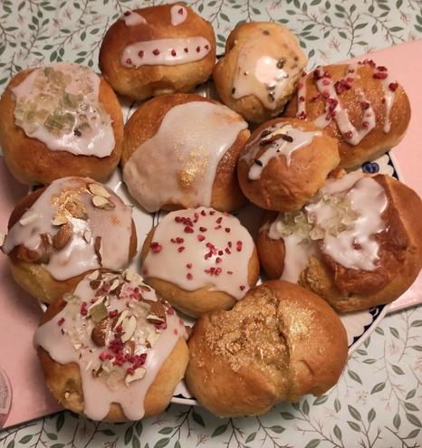 11 bread rolls on a plate, all lightly browned on top but now decorated with white icing and different sprinkles on top including freeze dried raspberries, chopped almonds, green sukat and a dusting of gold dust. All
