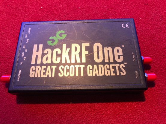 A black, rectangular plastic enclosure with the words "HackRF One" and "Great Scott Gadgets" on it in yellow letters. An antenna input is on the left side, and a clock input and output is on the right side, protected by cylindrical, soft plastic covers.