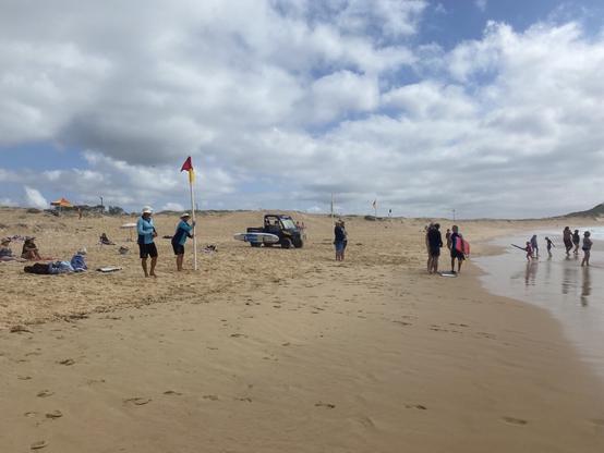 One surf lifesaver is adjusting the pole holding the red and yellow flag. The other is nearby watching the swimmers in the surf. They're both wearing a white hat, light blue shirt and shorts. People are sitting and lying on the beach. Others are walking along or playing at the water's edge.
