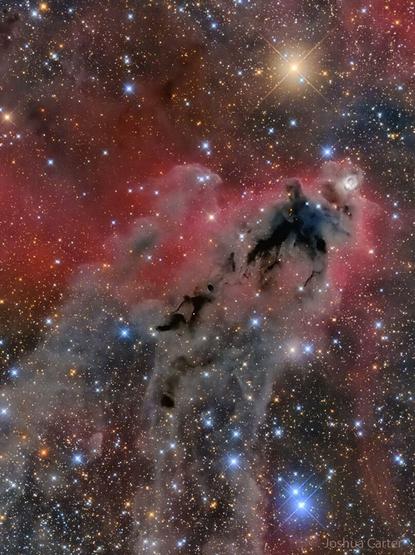 An image of a foreboding dark nebula before a red-glowing gas background and many bright and colorful stars.
