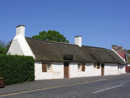 Burns Cottage in Alloway. The image shows a road in the foreground with a white cottage beyond the pavement on the far side. It has two brown doors and three windows, with brown shutters. The roof is thatched and there are two chimney stacks. The sky is blue.