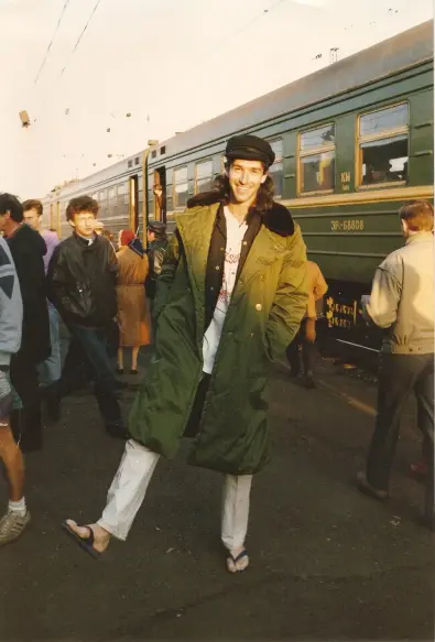 A young man with long dark hair under a black peaked cap is standing on front of a green train at a station platform. He is wearing a green greatcoat over a black waistcoat, white t-shirt, and slacks. He is standing with one foot slightly off the floor, possibly to draw attention to the fact that he is wearing sandals.