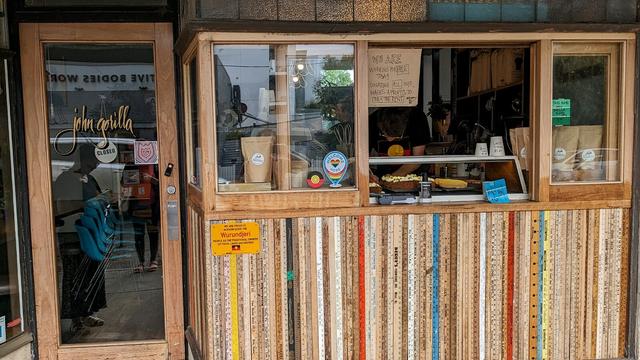 A photo of a small cafe. The front door is closed (with a "closed") sign, but the side serving window is open and two staff in Aboriginal flag t-shirts are making coffees and serving customers. The tip has has changed to a "pay the rent" collection, and a sign above the service window reads: "We are working for free today. Donating all our wages and profits to Pay the Rent." A permanent sign on the front of the building acknowledges the Wurundjeri people as the traditional owners of the land on which the cafe stands.